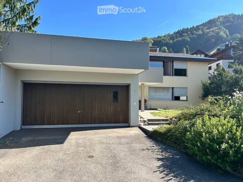 Modern two-story house with a large garage, clean lines, and a view of a green, hilly landscape in the background.