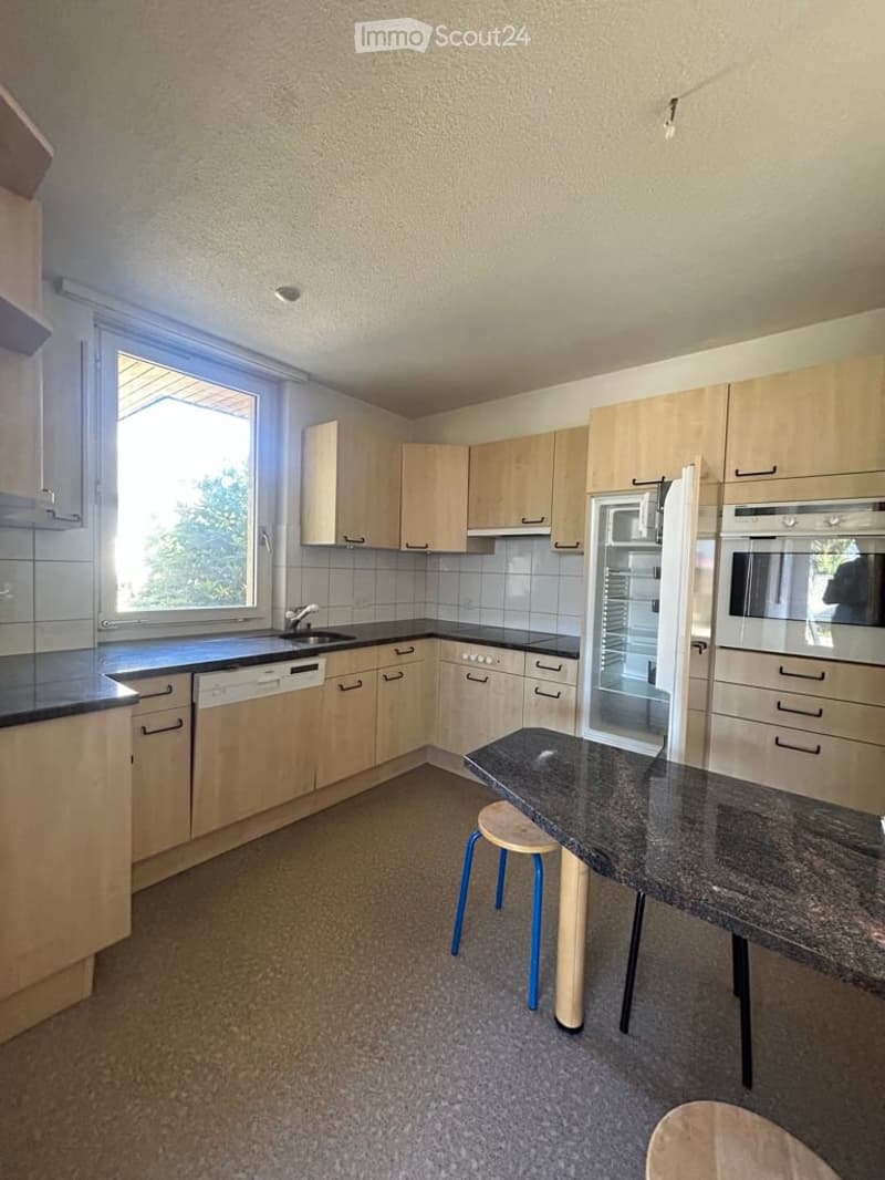 Modern kitchen with light wood cabinets, granite countertop, and built-in appliances including a refrigerator and dishwasher. A small table with stools is positioned near the window.