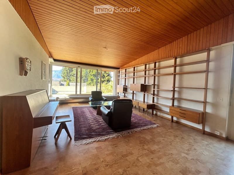Modern living room with wooden ceiling, large windows offering a scenic view, a purple area rug, a sofa, a chair, a glass coffee table, built-in wooden shelves, a small stool, and a piano.