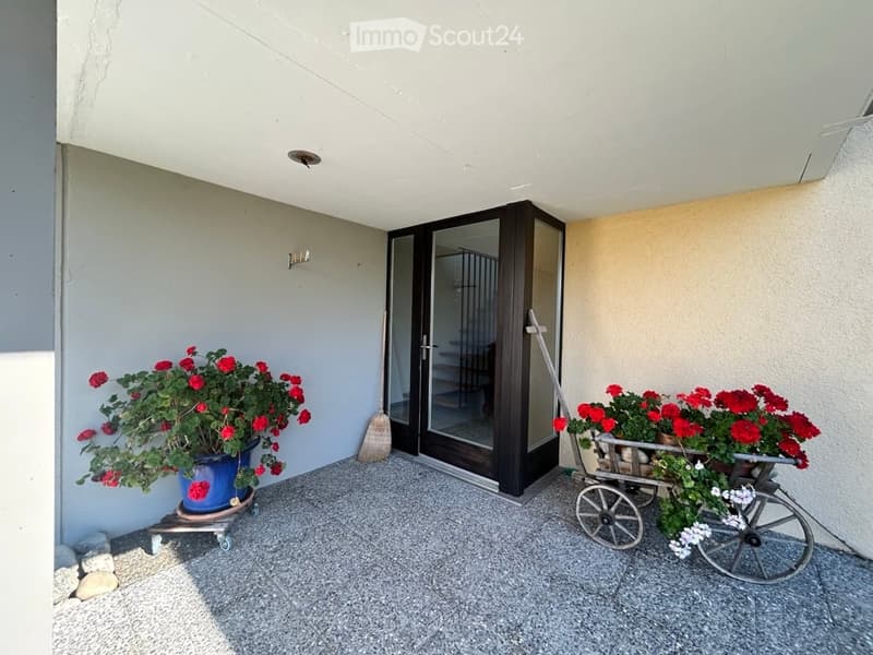 A small outdoor area with a covered entrance, potted plants, and a decorative cart with flowers.
