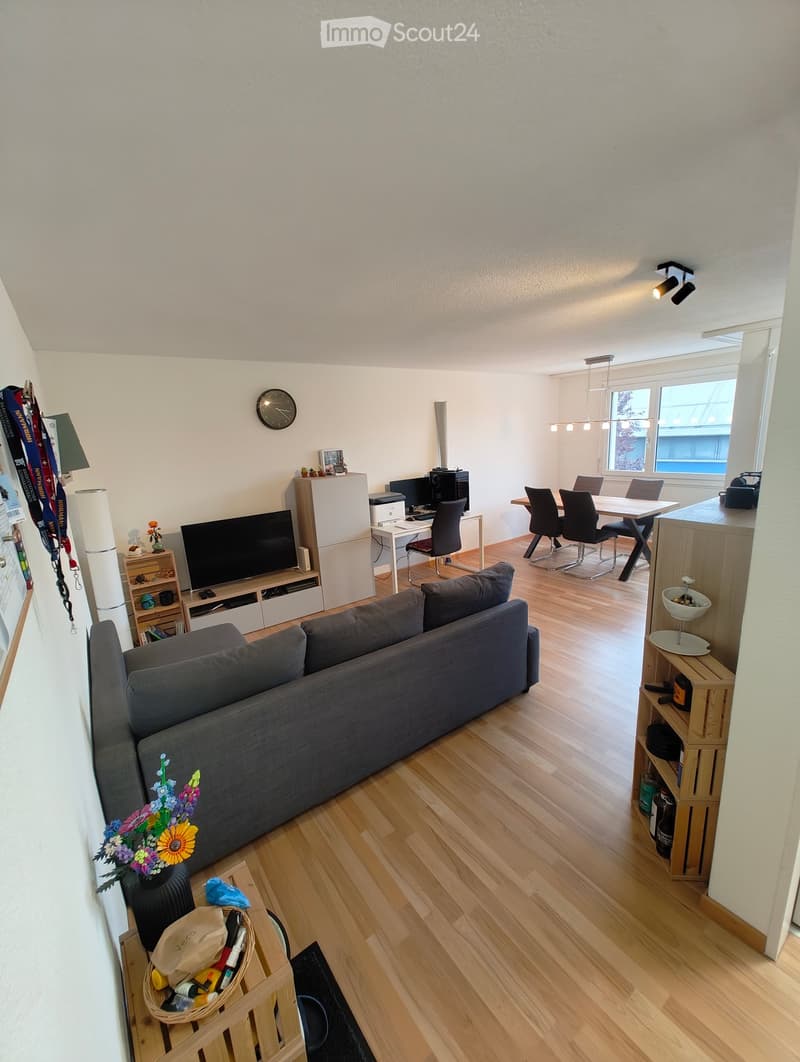 A modern living room with a grey sofa, a dining table with chairs, a refrigerator, a computer setup, and a TV stand with a screen. The room features wooden flooring and large windows with a view.