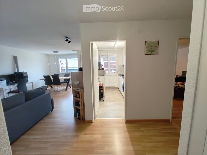 Bright, open-plan living and dining area with wooden flooring, a blue sofa, a dining table with chairs, a small kitchen area, and a bedroom visible in the background.