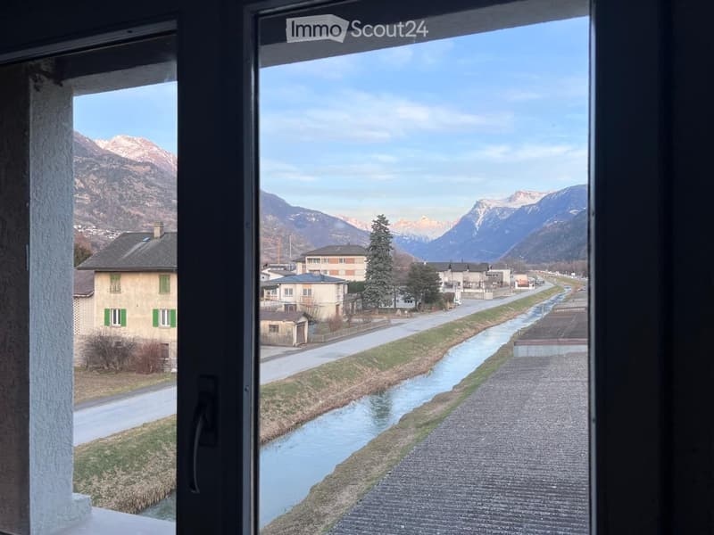 The image shows a scenic view from a window. It depicts a small town or village nestled in a mountainous landscape, with a river or stream running through the center. The buildings have a traditional European architectural style, with green shutters and r