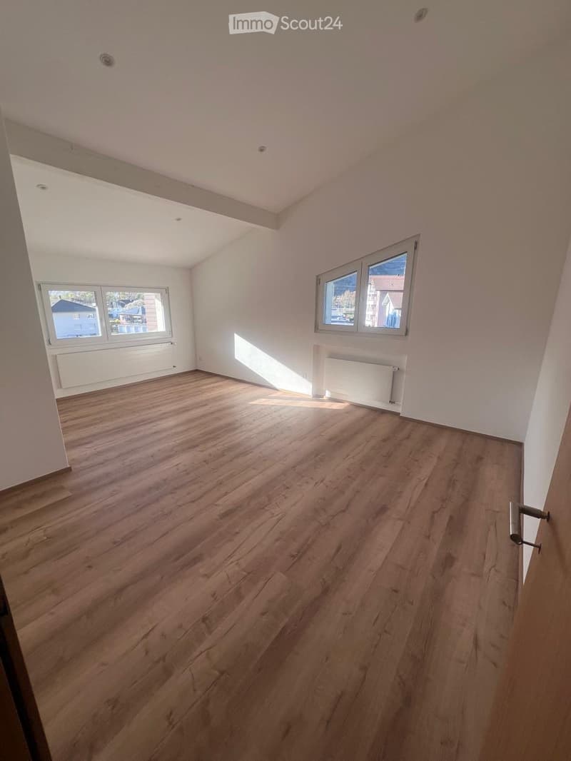 Spacious room with hardwood flooring, white walls, and large windows providing natural light. The room appears to be a living or bedroom area.