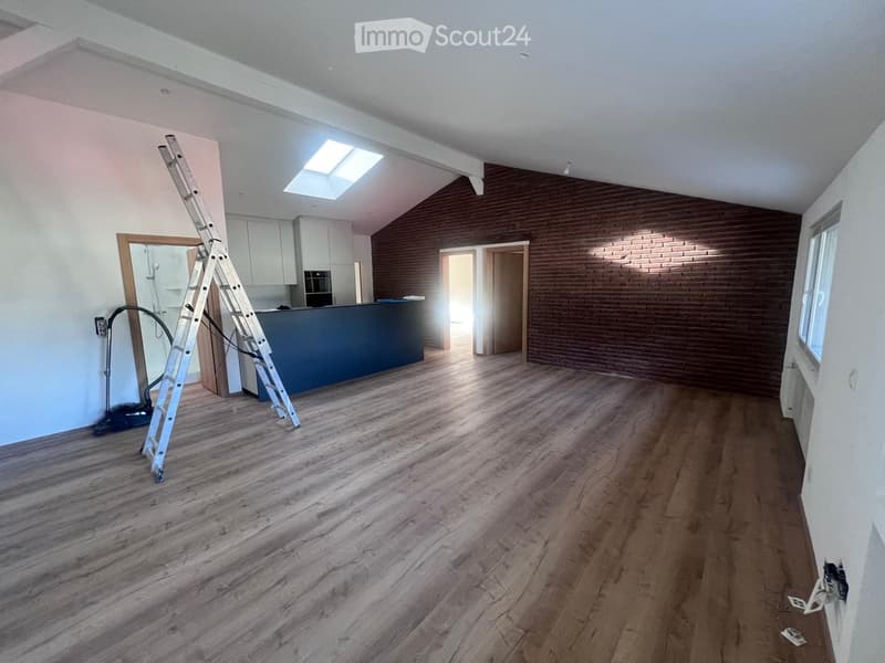 Open-concept living space with hardwood flooring, exposed brick wall, and a ladder leaning against the wall. The room has a kitchen area with a blue counter and appliances visible.