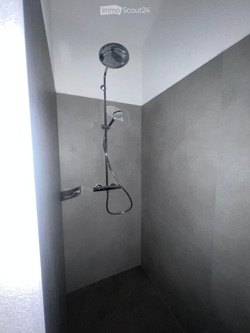 The image shows a bathroom with a shower head and faucet installed on the wall. The walls are a light gray color and the floor appears to be a dark tile or stone material.