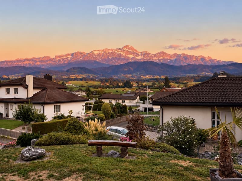 A peaceful garden area with a wooden bench, surrounded by greenery and flowers, with a car parked nearby. In the background, there are houses and a stunning mountain range under a clear sky.