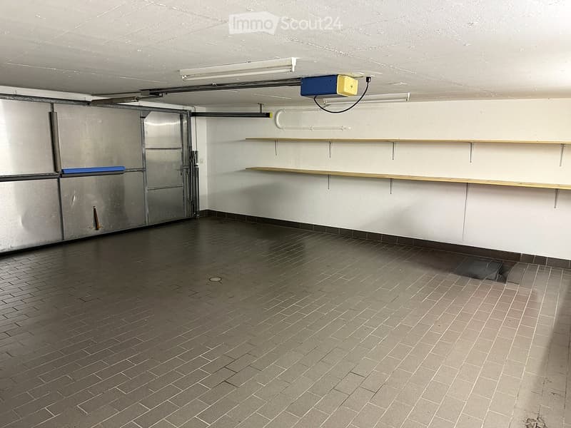 Empty room with tiled floor, white walls, and a metal door. There are wooden shelves mounted on the wall and a small electrical box on the ceiling.