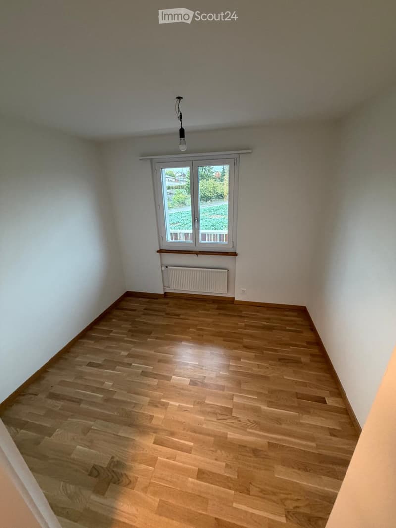 Empty room with wooden flooring, white walls, a window with a view of greenery, a radiator, and a ceiling light fixture.