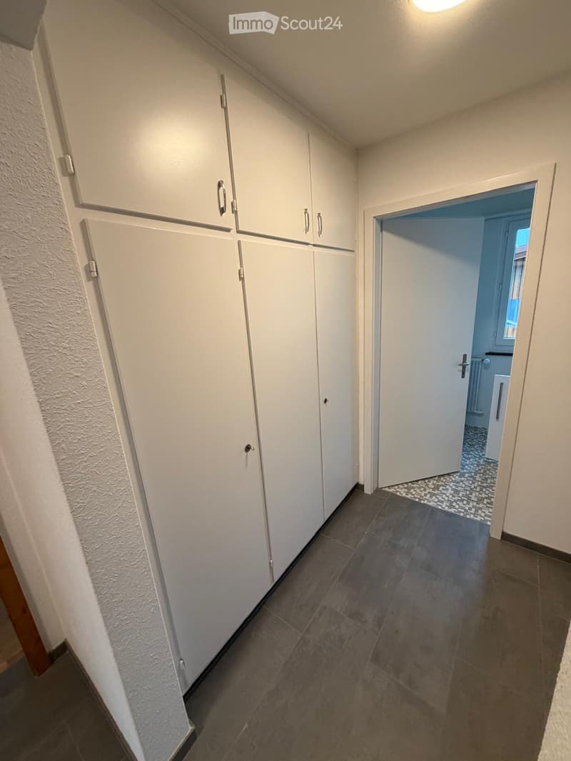 A narrow hallway with white built-in cabinets on one side and a tiled floor. An open doorway leads to a blue-tiled bathroom area visible in the background.