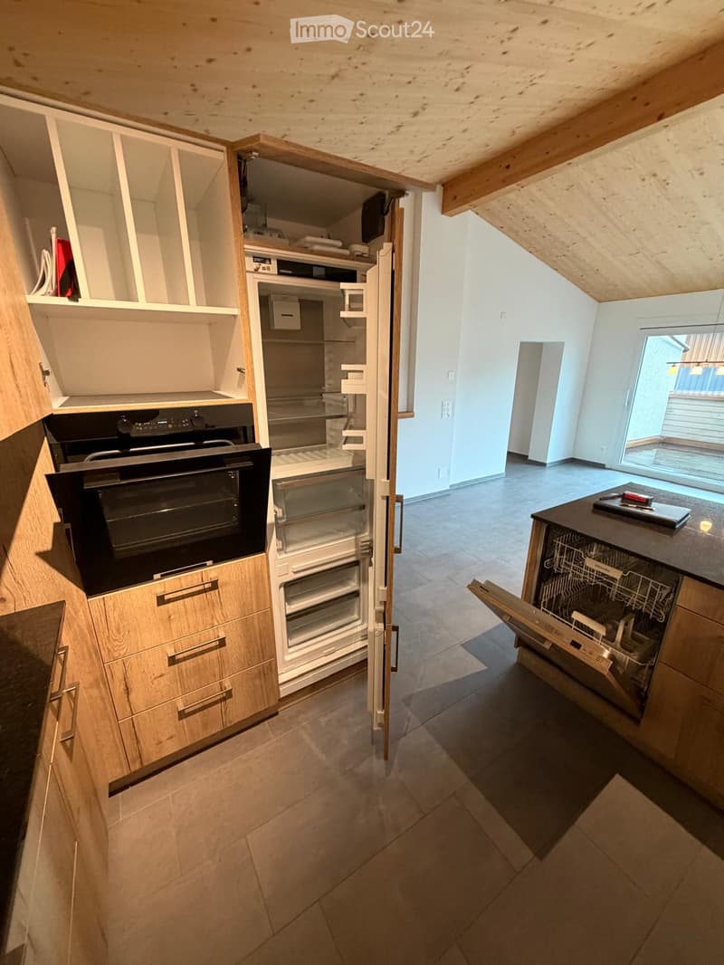 Modern kitchen with light wooden cabinets, stainless steel appliances including an oven, refrigerator, and dishwasher. The space features tiled flooring and an open layout with visible storage compartments.