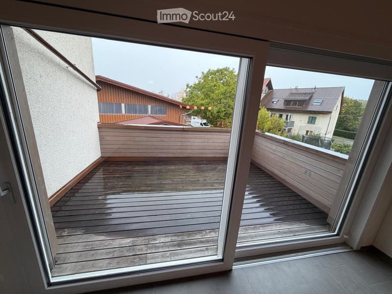 Wooden balcony with glass doors, overlooking a residential area with houses and trees.