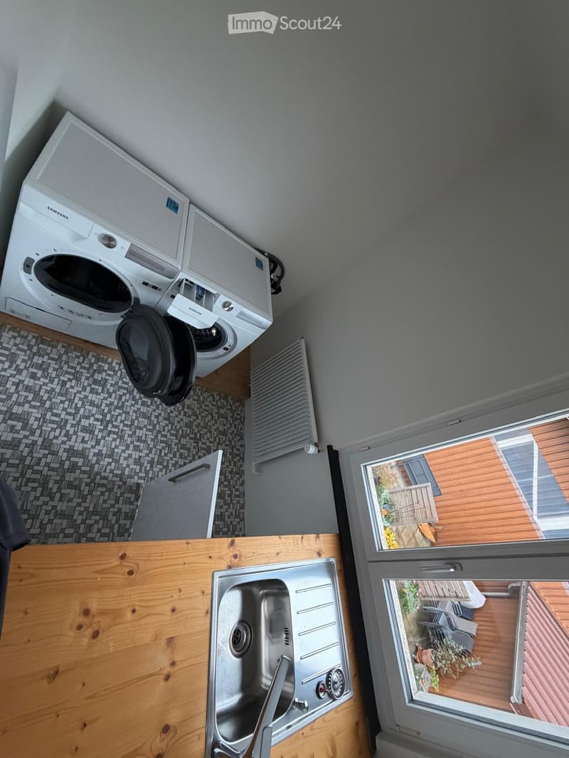 Bathroom with a stainless steel sink, wooden countertop, washing machine, radiator, and a window overlooking a wooden deck with outdoor furniture.