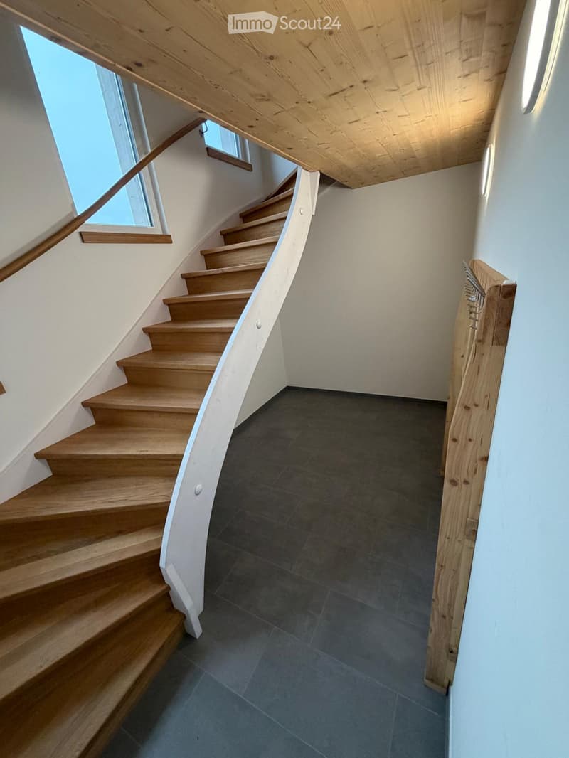 Wooden staircase with a modern design, featuring a sleek wooden handrail and a dark tiled floor. The walls are painted white, and the ceiling has exposed wooden beams.