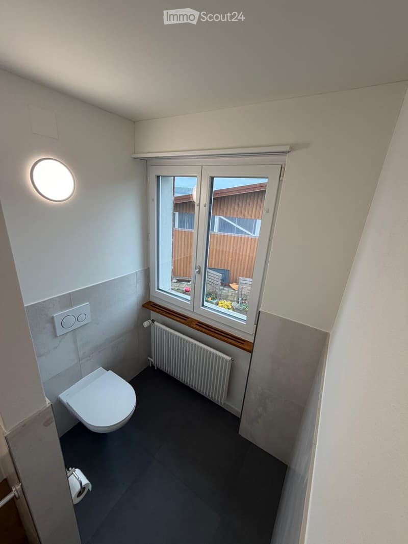 Small bathroom with a toilet, radiator, and a window offering a view of the garden. The room features modern tiles and a sleek design.