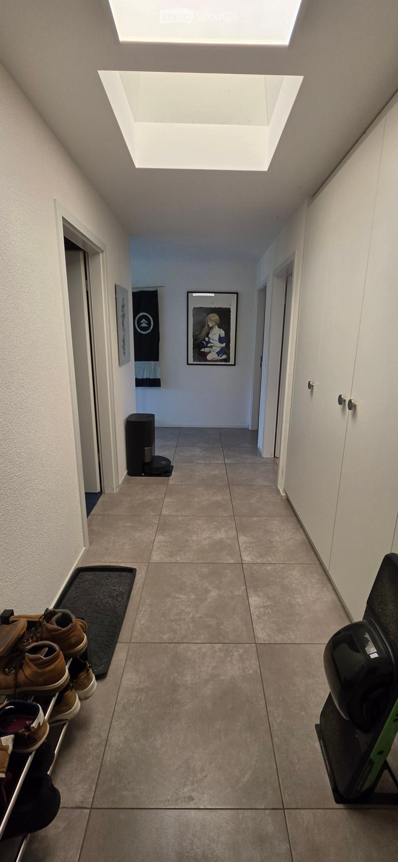 Clean, modern hallway with tiled floor, white walls, and a skylight at the top. Features include a shoe rack with shoes, a small black speaker, and artwork on the walls.