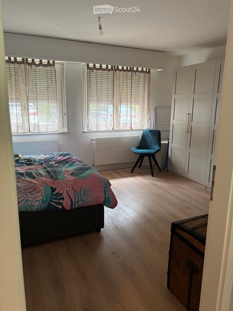 Bright bedroom with wooden flooring, large windows with blinds, a bed with a colorful bedspread, a modern blue chair, and a white wardrobe.