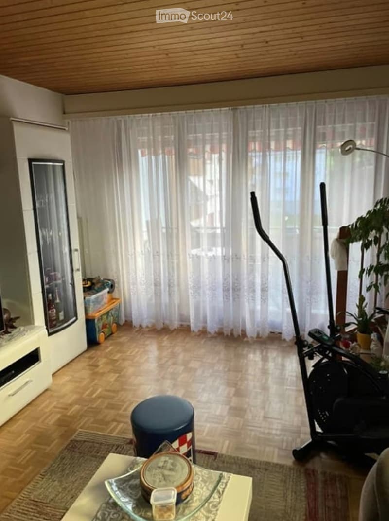 Living area with white curtains, wooden floor, table with a clock, stool, exercise bike, fridge, cabinet, and storage boxes.