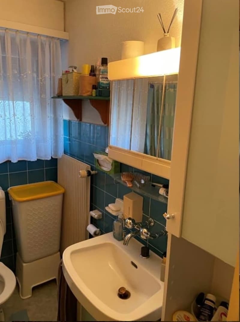 Bathroom with toilet, sink, mirror, shelves, radiator, trash can, curtains, and a yellow lid container.