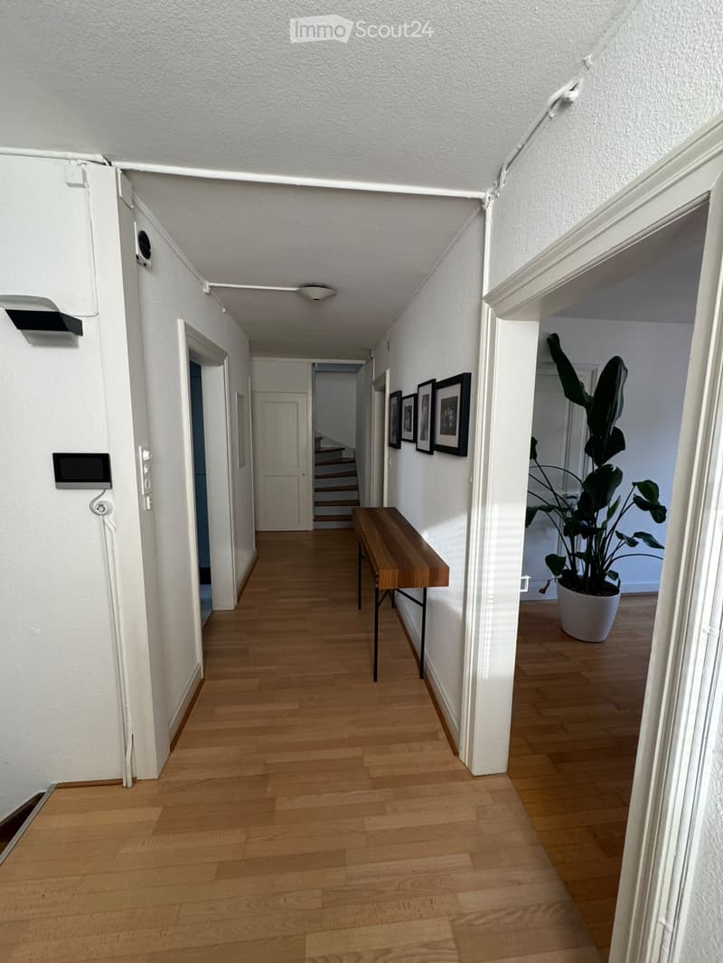 A well-lit hallway with wooden flooring, white walls, and a small wooden table placed against the wall. A large potted plant is visible in an adjacent room, and several framed pictures are displayed on the wall.