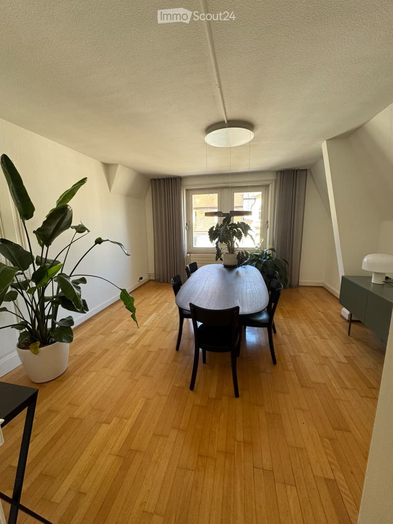 Dining room with wooden floor, large round dining table surrounded by chairs, potted plants, side table with lamp, and large window with curtains.