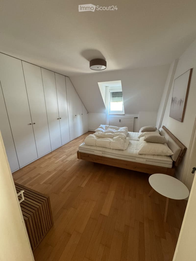 Cozy attic bedroom with wooden flooring, a bed with pillows and blankets, a small round table, and built-in white wardrobes.