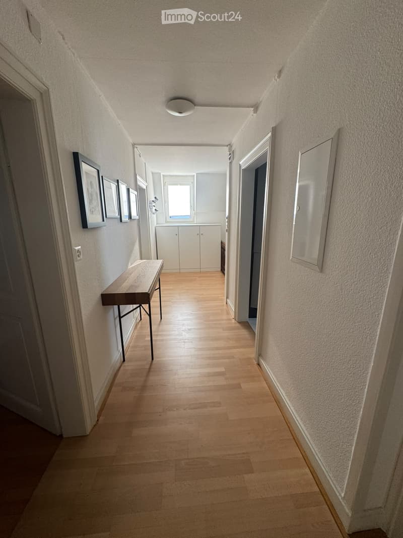 A narrow hallway with wooden flooring, white walls, and a small wooden table on the left side. There are framed pictures on the wall and a white cabinet at the end of the hallway.