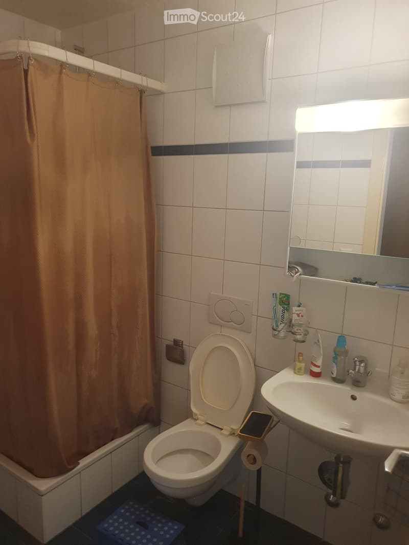 A compact bathroom with a toilet, sink, mirror, and shower area. The walls are tiled in white with a blue accent stripe. A shower curtain is visible, and various toiletries are placed on the sink counter.