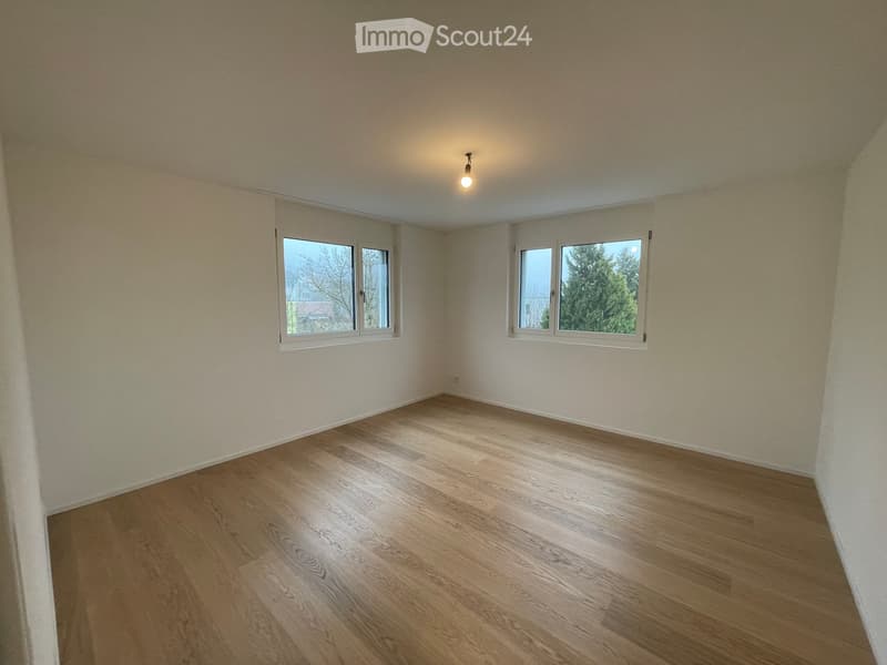 Empty room with light-colored walls, wooden flooring, and two windows offering a view of trees and a neighboring building.