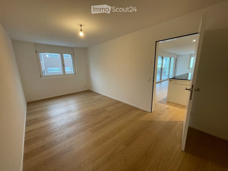 Empty room with wooden flooring, white walls, and a single window. A doorway leads to another room with a visible kitchen area.