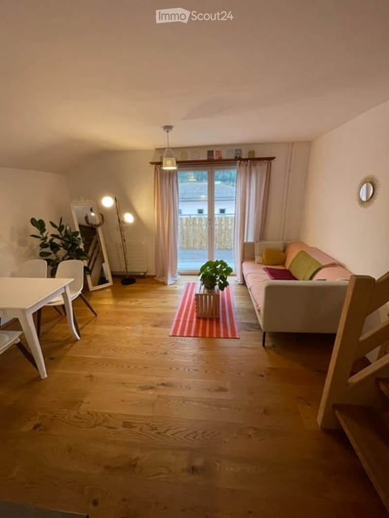 Bright living room with wooden flooring, large sliding glass door leading to a balcony, white sofa with colorful cushions, coffee table with a plant, dining area with white table and chairs, and a large mirror on the wall.