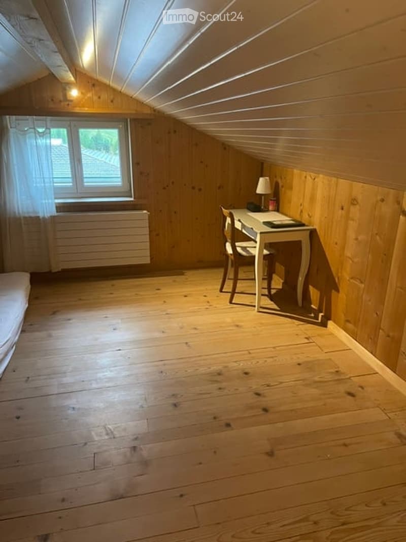 Cozy attic room with wooden walls and floor, featuring a small desk, chair, and lamp. Large window with white curtains allows natural light to fill the space.
