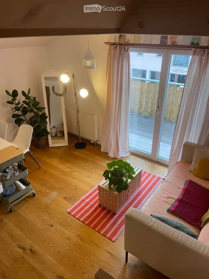 Bright living room with wooden flooring, large window and sliding glass door leading to a balcony, sofa, coffee table, chair, mirror, and plants.