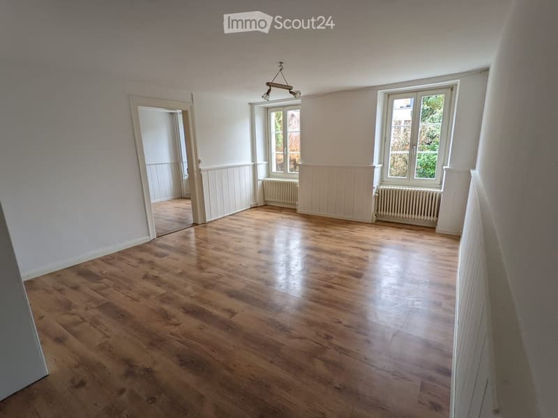 Empty room with wooden flooring, white walls, and large windows. There are radiators under the windows and a visible doorway leading to another space.