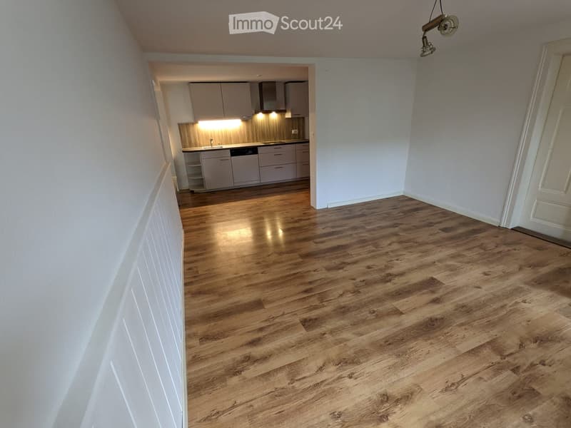 Empty room with wooden flooring and white walls, featuring an open doorway leading to a modern kitchen area with cabinets and appliances.