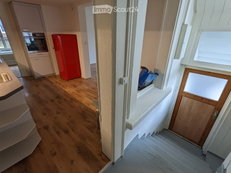 Hallway with wooden flooring, leading to a kitchen area with cabinets, a red refrigerator, and an oven. A staircase is visible on the right side.