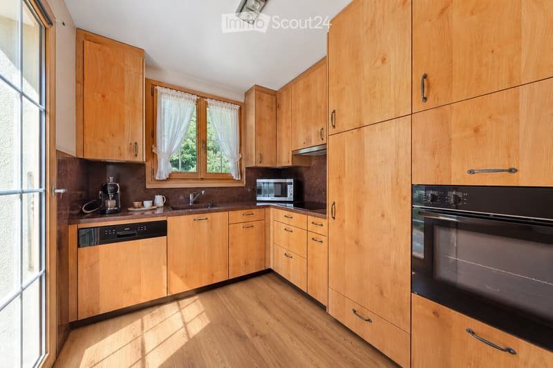 Bright kitchen with wooden cabinets, stainless steel appliances, and a window with white curtains. Includes a coffee maker, microwave, oven, dishwasher, and sink.