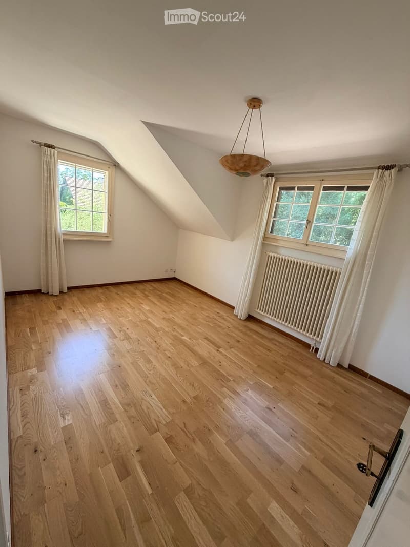 Empty attic room with wooden flooring, white walls, large windows with curtains, and a radiator. The ceiling has a sloped design with exposed beams.