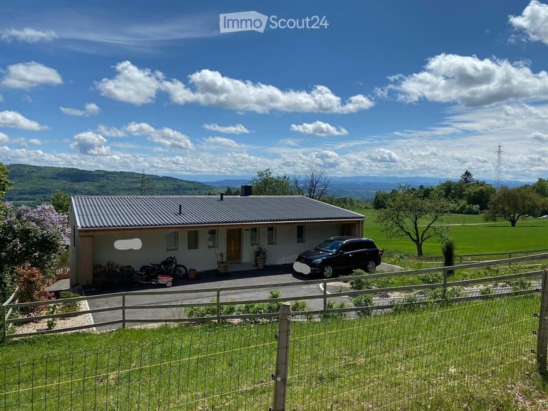 Single-story house with a metal roof, surrounded by a wooden fence, with a parked SUV and a motorcycle in the driveway, and a scenic rural landscape in the background.