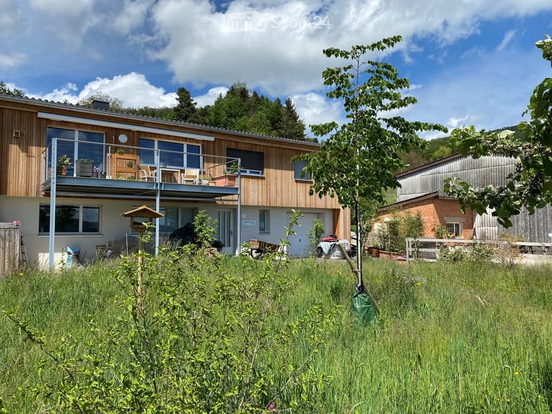Modern two-story house with wooden facade, large windows, and a balcony. The surrounding area includes a grassy yard, trees, and a small garden with outdoor furniture.