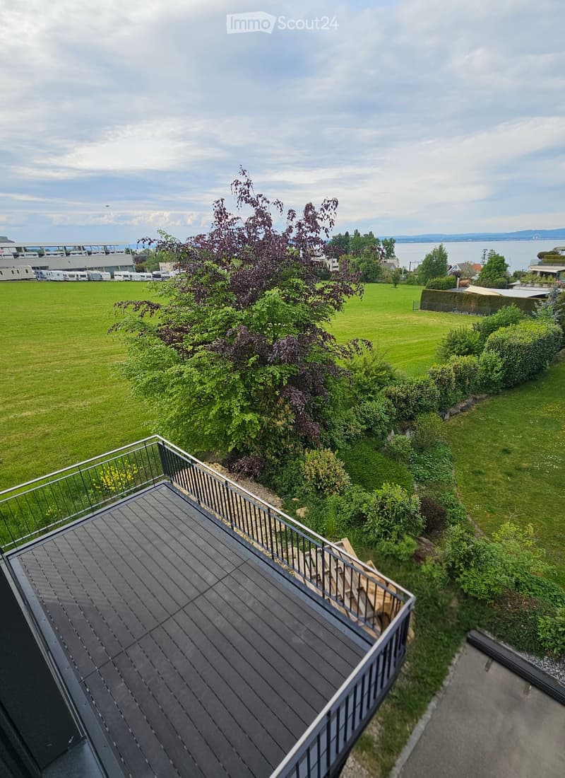 Wooden balcony with metal railing, overlooking a green lawn and distant water body.