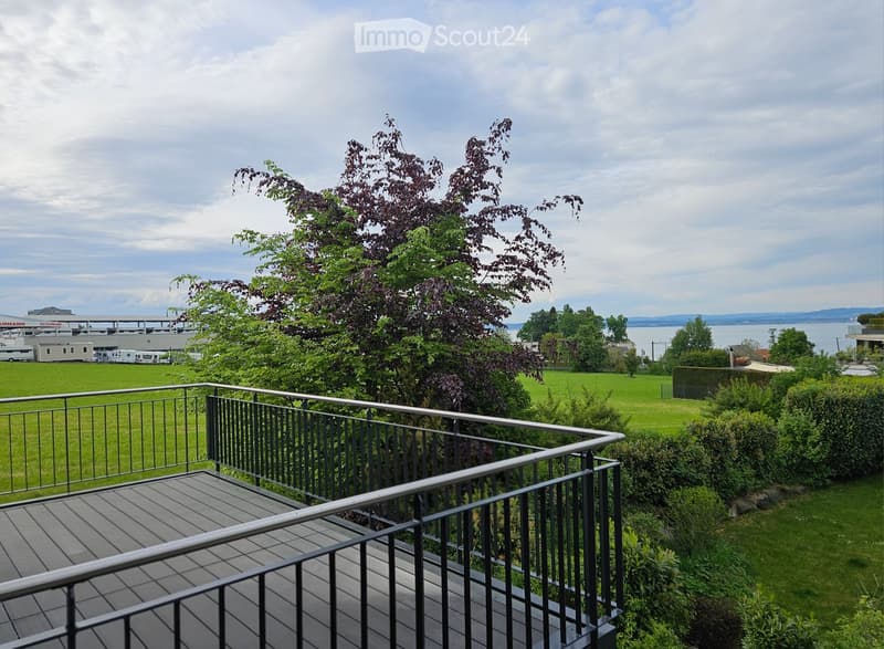 Balcony with wooden decking and metal railing, overlooking a green lawn, trees, and a body of water in the distance.