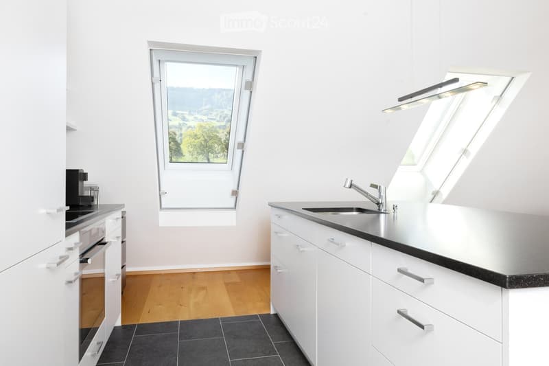 Modern kitchen with white cabinets, black countertops, stainless steel sink, and a coffee maker on the counter. Large windows offer a scenic view.