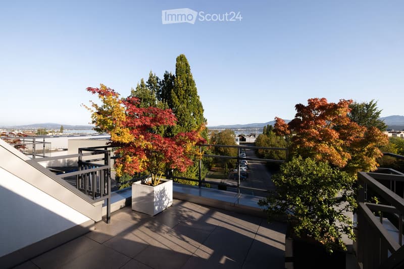 Modern balcony with tiled flooring, metal railing, and potted plants, overlooking a cityscape with trees and parked cars.