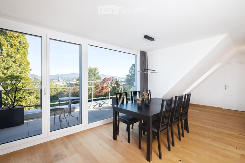 Modern dining room with wooden flooring, large windows, and a balcony overlooking a scenic view. The room features a black dining table with six chairs and a small table on the balcony.