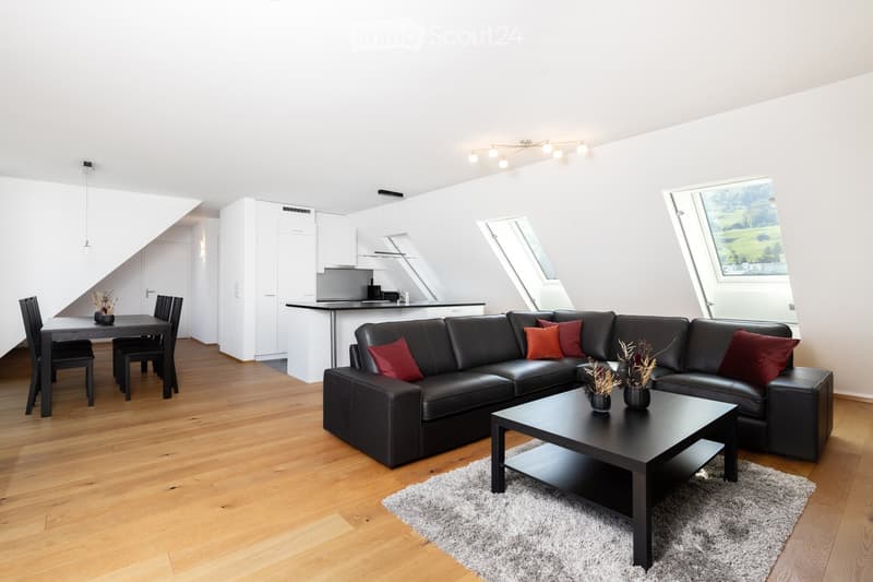 Modern living and dining area with a black leather sectional sofa, coffee table, dining table, and chairs. The space features wooden flooring, white walls, and large windows with a scenic view.