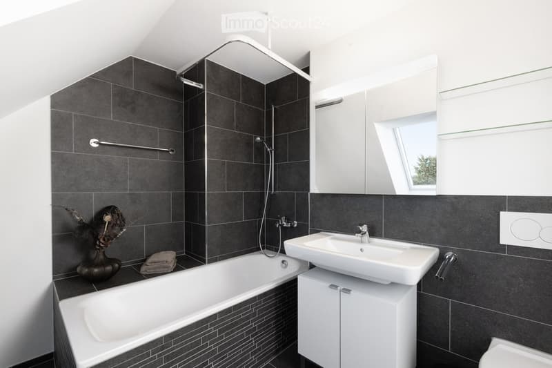 Modern bathroom with dark grey tiles, white bathtub, sink, mirror, and a small window letting in natural light.