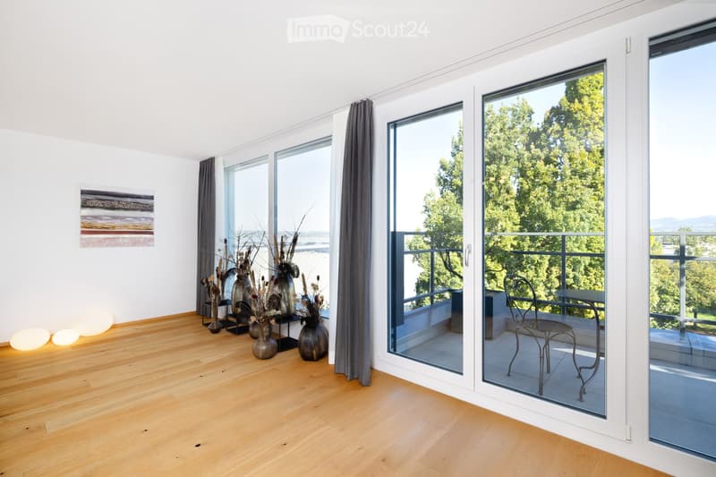 Bright living room with wooden flooring, large windows, and a balcony overlooking greenery. Decor includes vases, a chair, and a small table on the balcony.