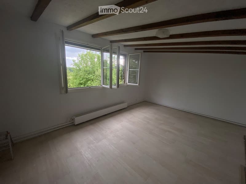 Empty room with wooden flooring, exposed wooden beams on the ceiling, and large windows offering a view of greenery outside.
