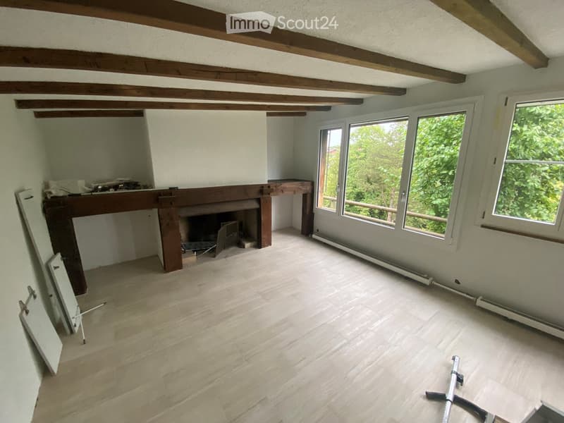 Empty room with wooden beams on the ceiling, light-colored wooden flooring, and a fireplace with a wooden mantel. Large windows provide natural light.